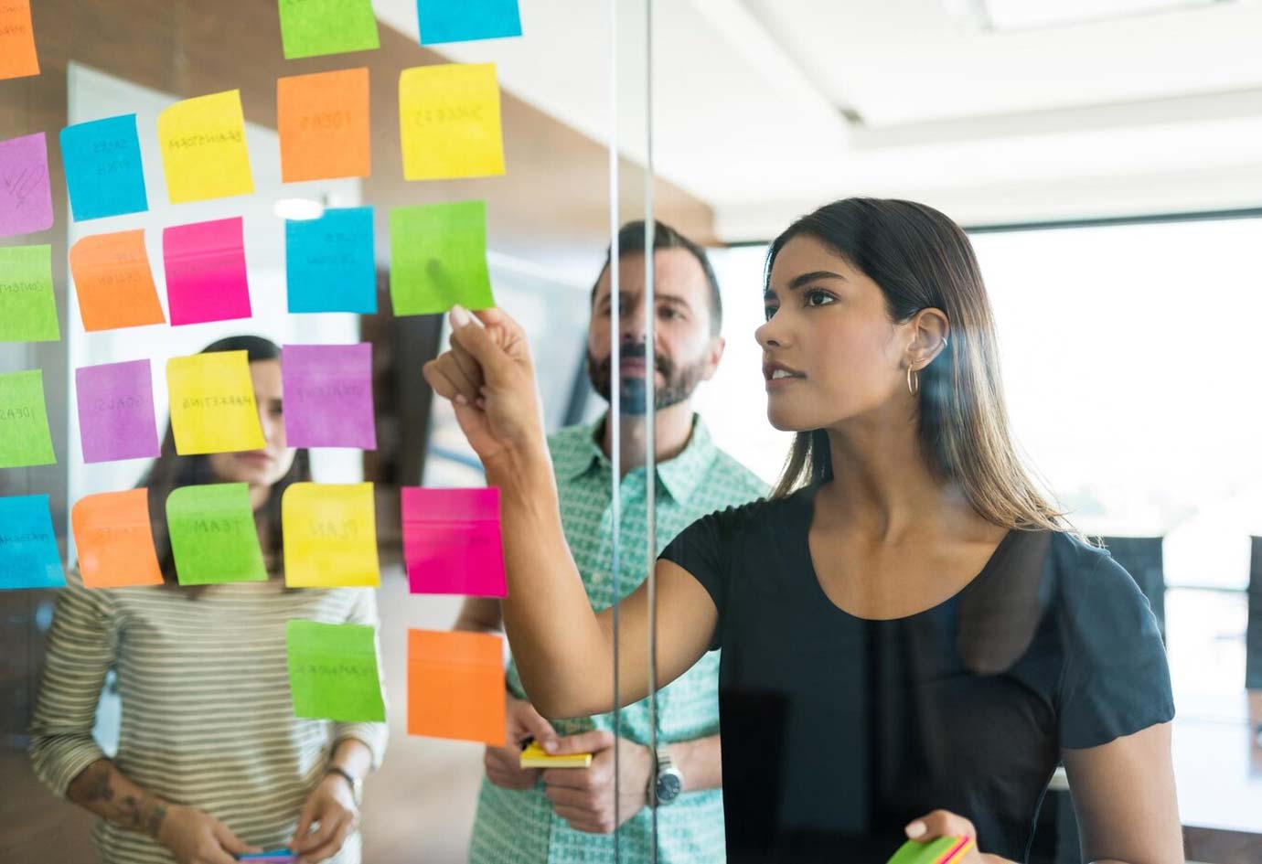 A photo of a woman sticking post-its on a wall of glass, explaining a plan for other employees